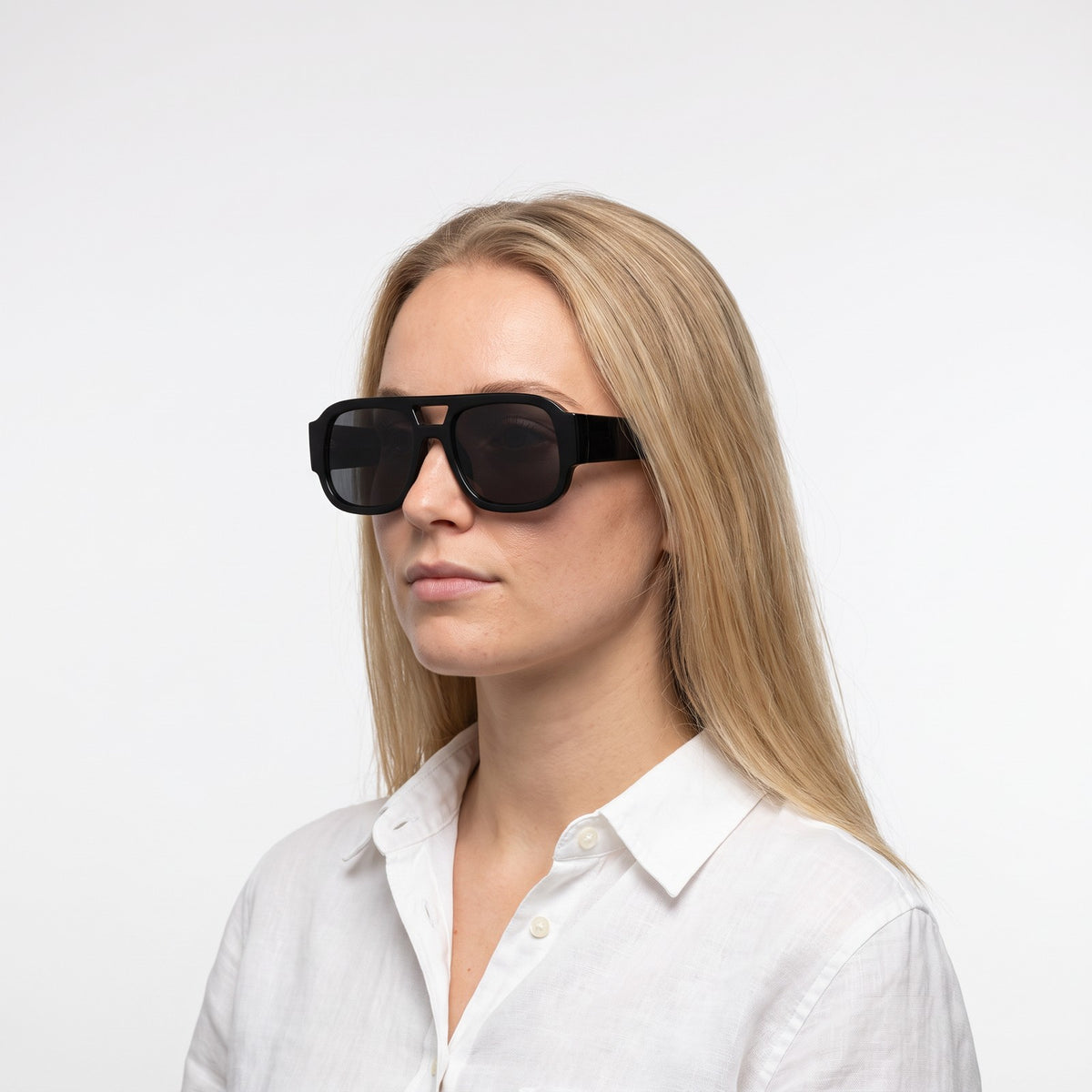 Woman in a white linen shirt wearing Crimson oversized square aviator sunglasses in Black with smoke lenses - Velezeco #opt1_2