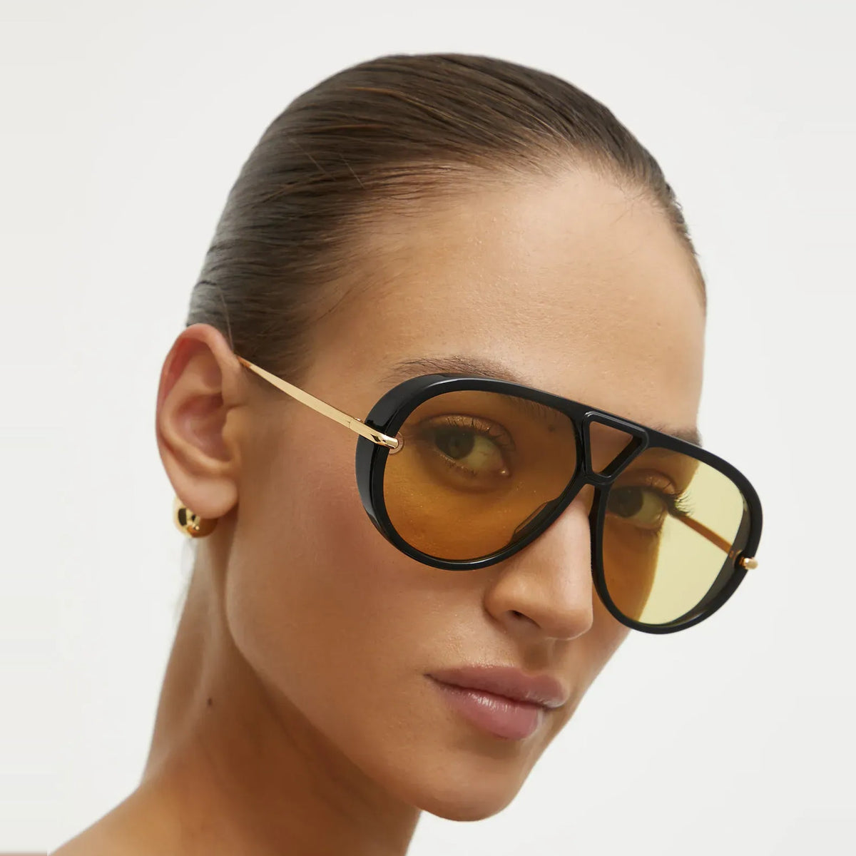 Close-up of The Nimbus oversized black aviator sunglasses with yellow tinted lenses and gold temples on a model, studio portrait — Velezeco #opt1_1